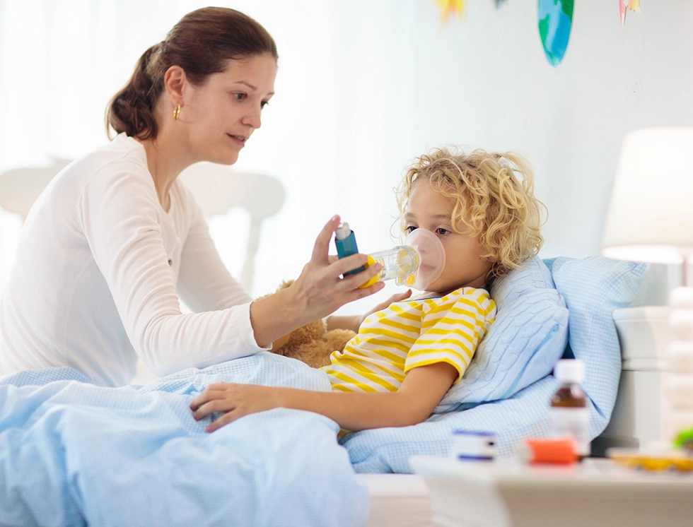 A caregiver administers medication to a child in bed, surrounded by soft blankets and a teddy bear, in a bright, cheerful room.