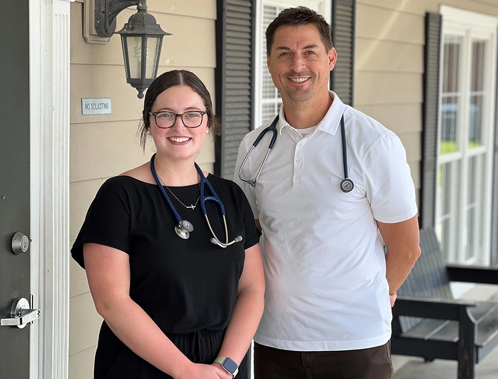 Two healthcare professionals, one in a black outfit with a stethoscope and the other in a white polo, stand outside a building.